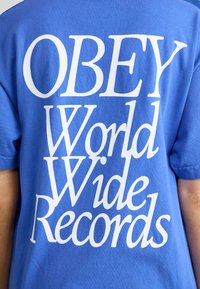Zila kokvilnas T-krekls ar lieliem baltiem uzrakstiem: "OBEY World Wide Records." Īsas piedurknes un brīva piegriezuma.