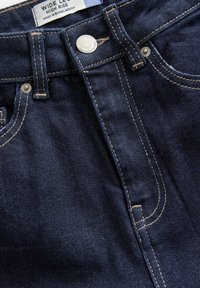 Vaqueros azul oscuro de talle alto y pierna ancha, con costuras visibles, botón de metal, remaches, trabillas para cinturón y etiqueta que dice "Wide Leg High Rise Made in Bangladesh."