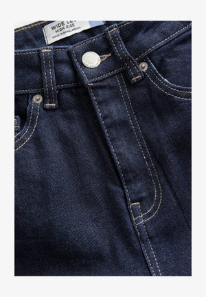 Wide-leg, high-rise dark denim jeans featuring visible stitching, a metal button closure, and a label indicating made in Bangladesh.