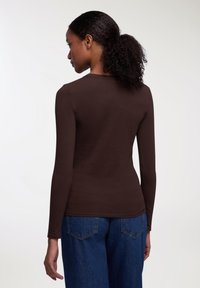 Camicia marrone a maniche lunghe con un design aderente, texture morbida e scollatura rotonda, abbinata a jeans blu a vita alta, vista da dietro.