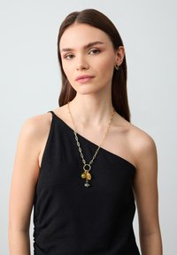Goldkette mit Anhängern, darunter ein Tannenzapfen und zwei Perlen, getragen über einem schwarzen einseitigen Oberteil mit gerafften Details.