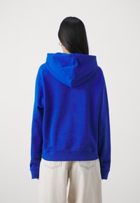 Niebieska bluza z kapturem o luźnym kroju, z prążkowanymi mankietami i dołem. Gładka tekstura materiału, widoczne przeszycia na ramionach i u dołu.