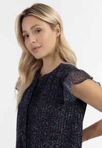 Blusa floreale navy con maniche trasparenti arricciate, parte frontale plissettata e scollatura rotonda. Presenta motivi floreali colorati e texture sottili.