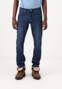 Hombre con jeans de mezclilla azul oscuro y botas de cuero marrón con puños de piel doblados, de pie contra un fondo blanco liso.