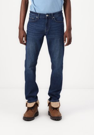 Hombre con jeans de mezclilla azul oscuro y botas de cuero marrón con puños de piel doblados, de pie contra un fondo blanco liso.