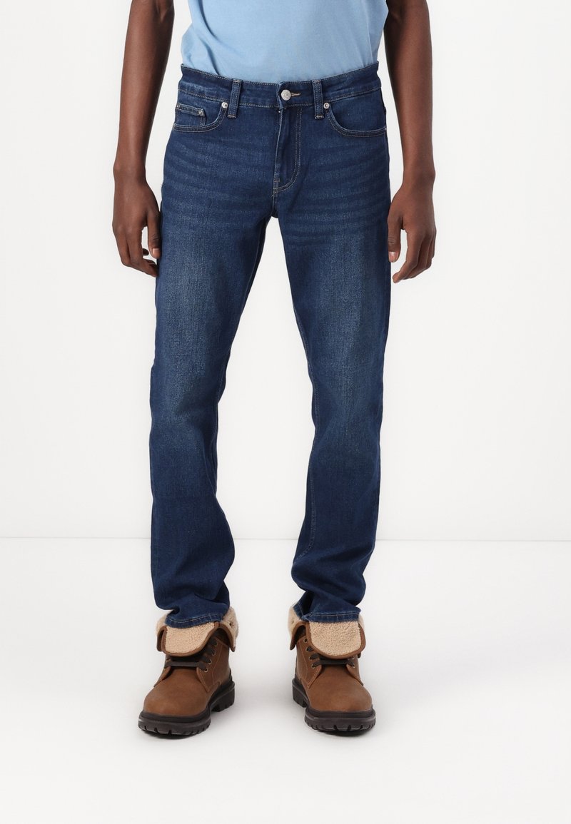 Hombre con jeans de mezclilla azul oscuro y botas de cuero marrón con puños de piel doblados, de pie contra un fondo blanco liso.