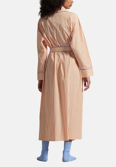 Długa, pomarańczowo-biała szata w paski z szerokimi rękawami i paskiem do przewiązania, ozdobiona kontrastową niebieską lamówką na mankietach. Modelka ma na sobie niebieskie, dziergane skarpety.