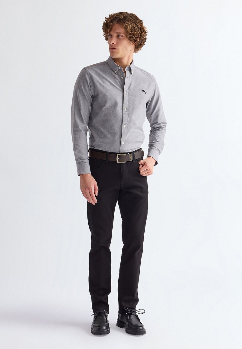 Junger Mann mit lockigem Haar, der ein hellgraues Hemd mit Knöpfen, schwarzen Gürtel, schwarze Hose und schwarze Schuhe trägt, steht vor einem weißen Hintergrund.