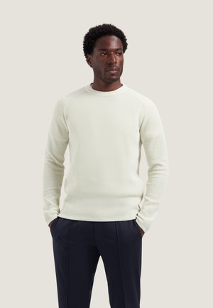 Homme portant un pull blanc texturé à manches longues, les mains dans les poches, associé à un pantalon bleu marine foncé, debout sur un fond clair.