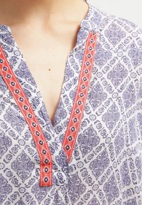 Krage och urringning av mönstrad blå och vit skjorta med röd dekorativ trim på en persons övre bröst och nacke.