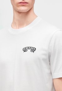 Jeune personne portant un t-shirt blanc avec un petit logo noir "DIESEL" sur la partie supérieure gauche de la poitrine.
