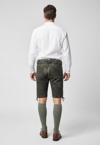 Mörkgröna lädershorts för män med sidoflätade detaljer, tillsammans med knälånga texturerade grå strumpor och bruna skor. Vit skjorta synlig.
