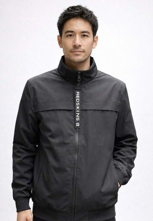 Homme portant une veste noire zippée avec "REDSKINS" sur la tirette de la fermeture éclair, debout devant un fond blanc uni.