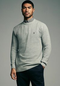 Lys grå strikket sweater med tekstureret design, ribbede manchetter og kant, med et lille sort logo på brystet, båret over en skjorte med krave.