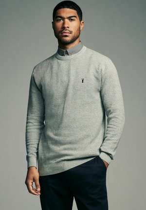 Lys grå strikket sweater med tekstureret design, ribbede manchetter og kant, med et lille sort logo på brystet, båret over en skjorte med krave.