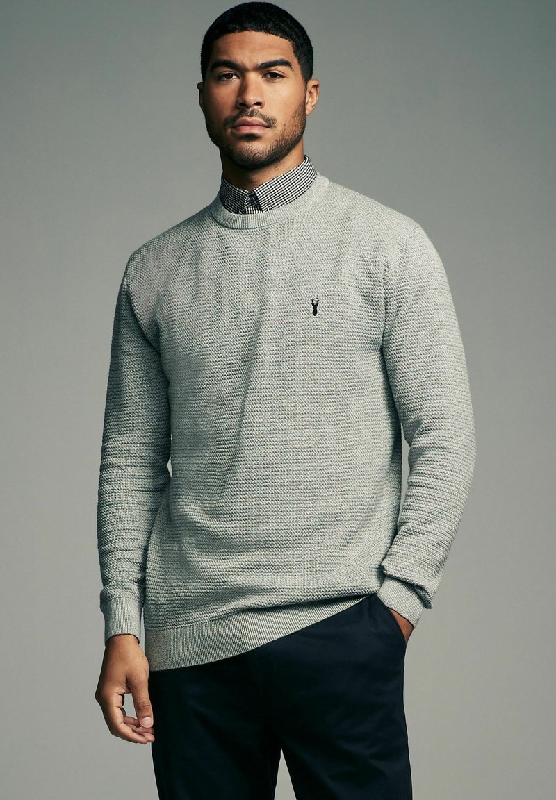 Lys grå strikket sweater med tekstureret design, ribbede manchetter og kant, med et lille sort logo på brystet, båret over en skjorte med krave.