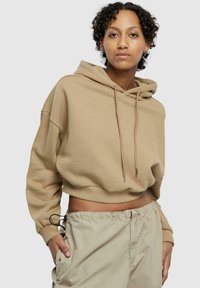 Avskåret beige hettegenser med snor, lange ermer og en myk tekstur. Brukt sammen med lyse beige bukser med sidelommer og elastisk midje.