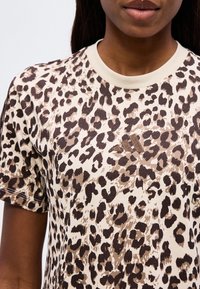 Femme portant un t-shirt beige à manches courtes avec un imprimé léopard marron et noir et un logo Adidas brodé discret sur la poitrine.