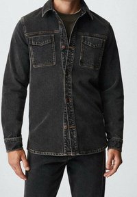 Chaqueta de mezclilla negra con dos bolsillos en el pecho, cierres de botón y costuras en contraste. Textura suave con un corte relajado y un cuello.