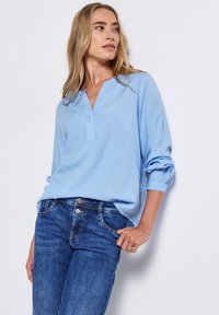 Blouse bleu clair avec un décolleté en V, des manches longues et des poignets froncés, associée à un jean bleu foncé. Le tissu semble lisse et léger.