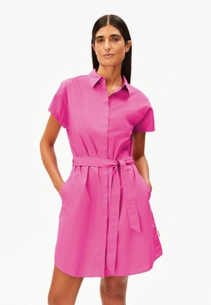 Femme portant une robe-chemise ceinturée à manches courtes de couleur rose vif avec col, debout les mains dans les poches, sur un fond blanc uni.