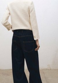 Chaqueta de crema texturizada con acentos de botones dorados, combinada con vaqueros de pierna ancha azul oscuro con dos bolsillos traseros y costuras en contraste.