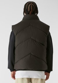 Zwarte puffer-vest met een hoge kraag, voorzien van een chevronpatroon dat in de stof is gestikt. Draagt over een zwart lang-sleeve shirt.