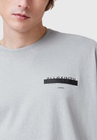 Camiseta de algodón gris con un diseño gráfico negro que incluye el texto "ALLSAINTS" y una franja negra horizontal en el centro.