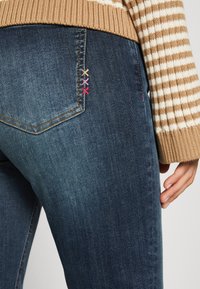 Vaqueros de mezclilla azul oscuro con bolsillos traseros en ángulo, que presentan acentos de costura multicolor. Textura suave con una silueta ajustada.