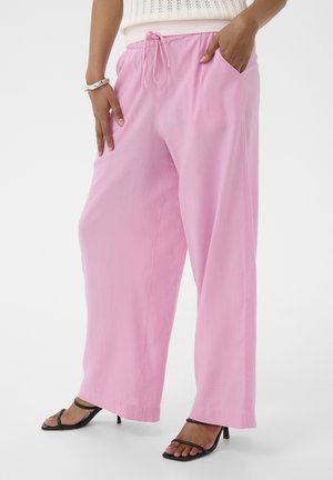 Nő laza, világos rózsaszín, zsinórderékpántos nadrágban és fekete, pántos, magassarkú szandálban, kezét a zsebében tartva, fehér háttér előtt áll.