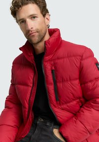 Crvena podstavljena jakna s visokim ovratnikom, horizontalnim šavovima, zatvaračima na rajsferšlus i crnim akcentima. Nosena preko crne majice, prikazana na neutralnoj pozadini.