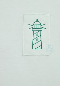 Baltas kokvilnas T-krekls ar zaļu izšūtu bākas dizainu taisnstūrveida plāksterī un gaiši zilu izšūtu burtiņu "R" akcentu.