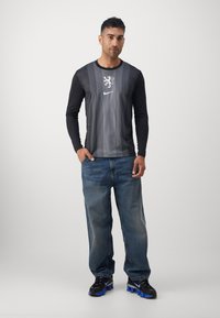 Langarmige zwart-grijs gestreepte shirt met leeuwlogo en Nike-branding, gecombineerd met blauwe jeans en zwarte sneakers met blauwe accenten.