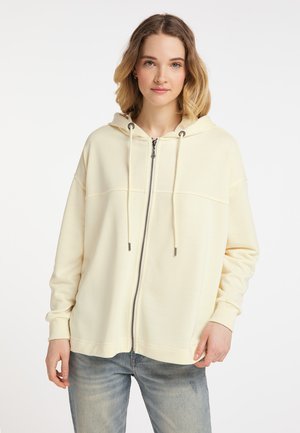 Jonge vrouw met een crème kleurige zip-up hoodie met trekkoorden en lichtblauwe jeans, staand tegen een effen witte achtergrond.