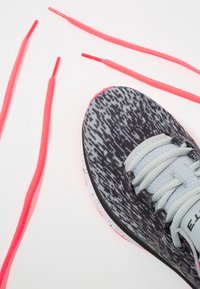 Scarpa sportiva con motivo nero e grigio, con lacci grigio chiaro e un accento rosa sulla suola. Vicino ci sono quattro lacci color rosa brillante.