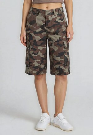 Mujer con pantalones cortos cargo de camuflaje hasta la rodilla y zapatillas blancas, de pie contra un fondo liso y claro.