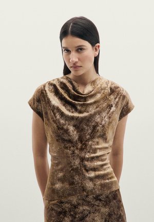 Femme aux cheveux lisses et foncés portant un haut en velours marron brillant avec un décolleté drapé sur un fond clair uni.