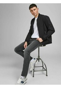 Black bomber jacket over a white t-shirt, gray jeans, and gray sneakers. Man seated on a metal stool with a neutral background.