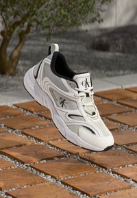 Biele športové topánky s sieťovinou, čiernymi akcentmi a textúrovanou gumovou podrážkou. Obsahujú logo Calvin Klein na jazyku a na boku.