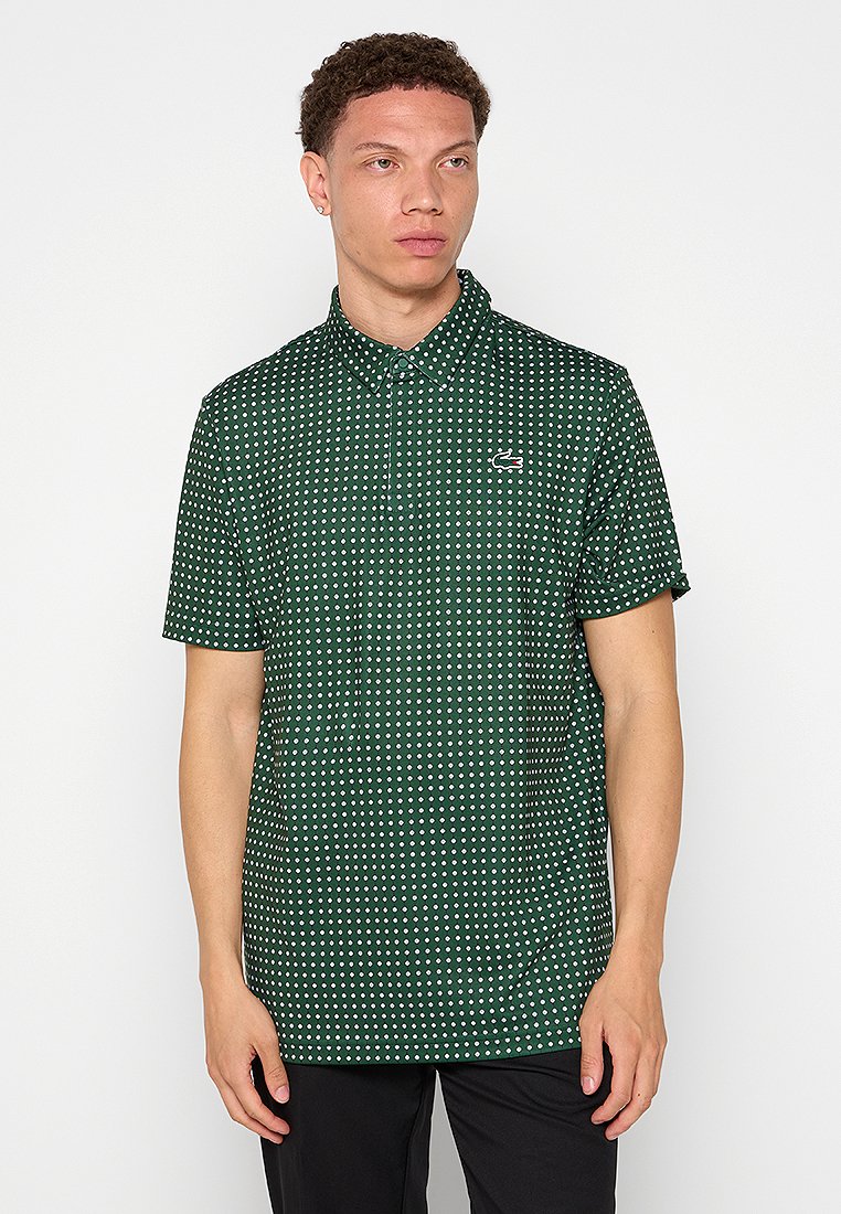 Joven vestido con una camiseta polo de manga corta verde, con lunares blancos y un pequeño logo en el pecho, de pie frente a un fondo blanco.