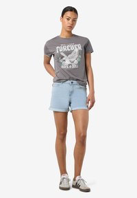 Žena stojaca v šedej tričku s grafikou "Forever Rock & Roll", svetlých modrých džínsových šortkách a bielych teniskách Adidas s čiernymi pruhmi.