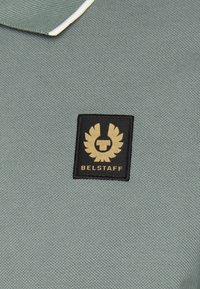 Zelená polo košile vyrobená z texturované látky, s černým čtvercovým nášivkem se zlatým logem a textem "BELSTAFF" ve středu.