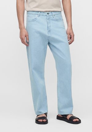 Person iført lyseblå jeans med lige ben, beige top og sorte sandaletter med åbne tæer, stående mod en ensfarvet baggrund.