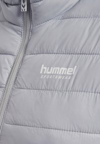 Hellgraue gesteppte Sportjacke mit vertikalem Reißverschluss und dem "hummel sportswear" Logo auf der Brust.