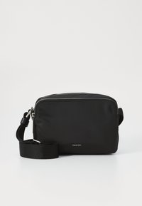 Geantă transversală mică din nylon negru, cu închidere cu fermoar, curea ajustabilă și accente din metal argintiu. Logo-ul Calvin Klein este afișat pe față.