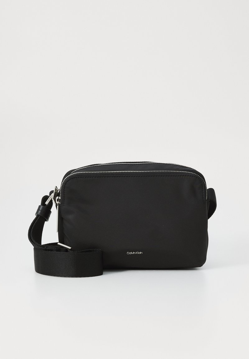 Geantă transversală mică din nylon negru, cu închidere cu fermoar, curea ajustabilă și accente din metal argintiu. Logo-ul Calvin Klein este afișat pe față.