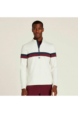 Uomo che indossa una maglia a maniche lunghe bianca con righe orizzontali blu navy e bordeaux e una mezza cerniera, in piedi davanti a uno sfondo semplice.