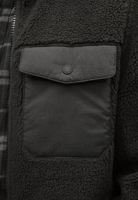 Fekete gyapjú pulóver texturált felülettel, sima anyagból készült lapos mellzsebbel, amelyet kör alakú patent díszít.