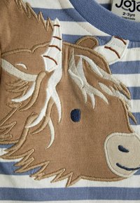 Tan pruhovaná košela s vyšitým dizajnom zvieracej hlavy, detailným kožuchom, modrými akcentmi a textúrovaným povrchom.