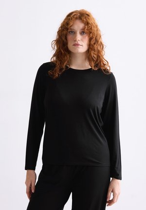 Rothaarige Frau mit lockigem Haar, die ein schlichtes schwarzes Langarmshirt und schwarze Hosen trägt, vor einem weißen Hintergrund.